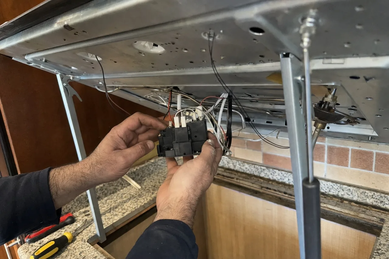 A gas cooktop lifted clear of the granite worktop and held open on supports while a technician holds the spark module and its wiring harness underneath