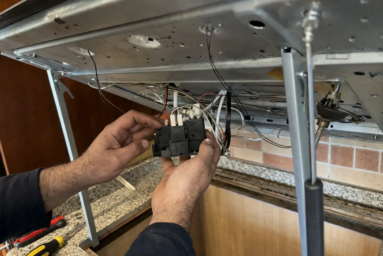 Bare hands holding a black ignition switch block and its loom under a lifted cooktop, with screwdrivers laid out on the counter behind