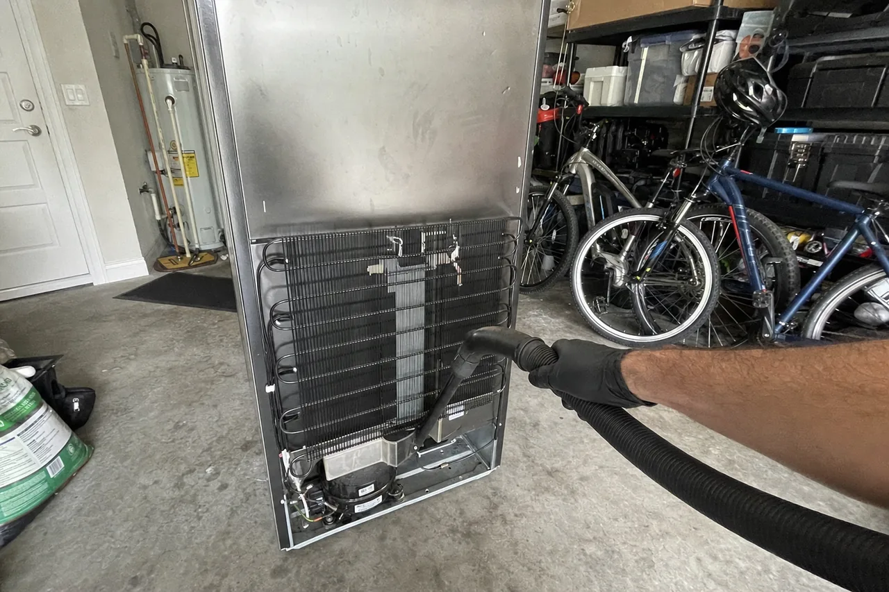 A refrigerator pulled out from the garage wall with its lower rear panel removed, the condenser coil being vacuumed clean