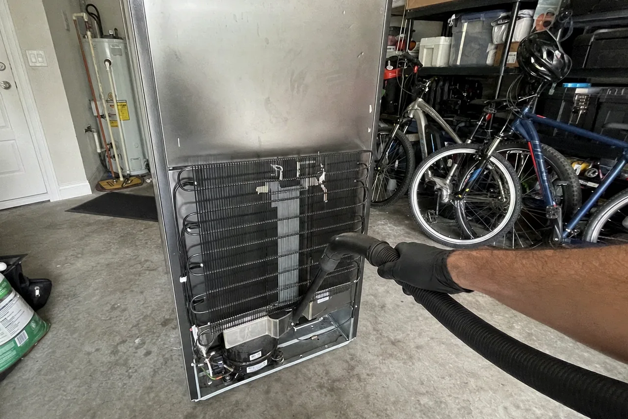A stainless refrigerator standing away from the wall in a home garage with its back service panel off and the coil exposed, bicycles racked behind it