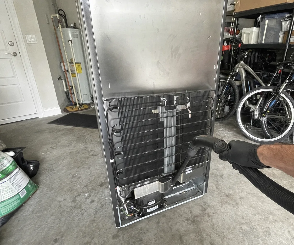 The condenser coil and machine compartment of a stainless refrigerator with the rear panel removed, a vacuum hose drawing dust off the fins