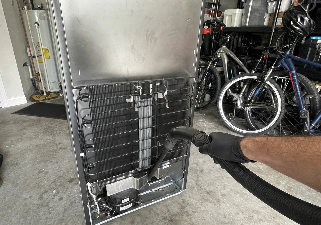 A stainless refrigerator wheeled out into the middle of a home garage for a coil clean, water heater and shelving along the wall behind