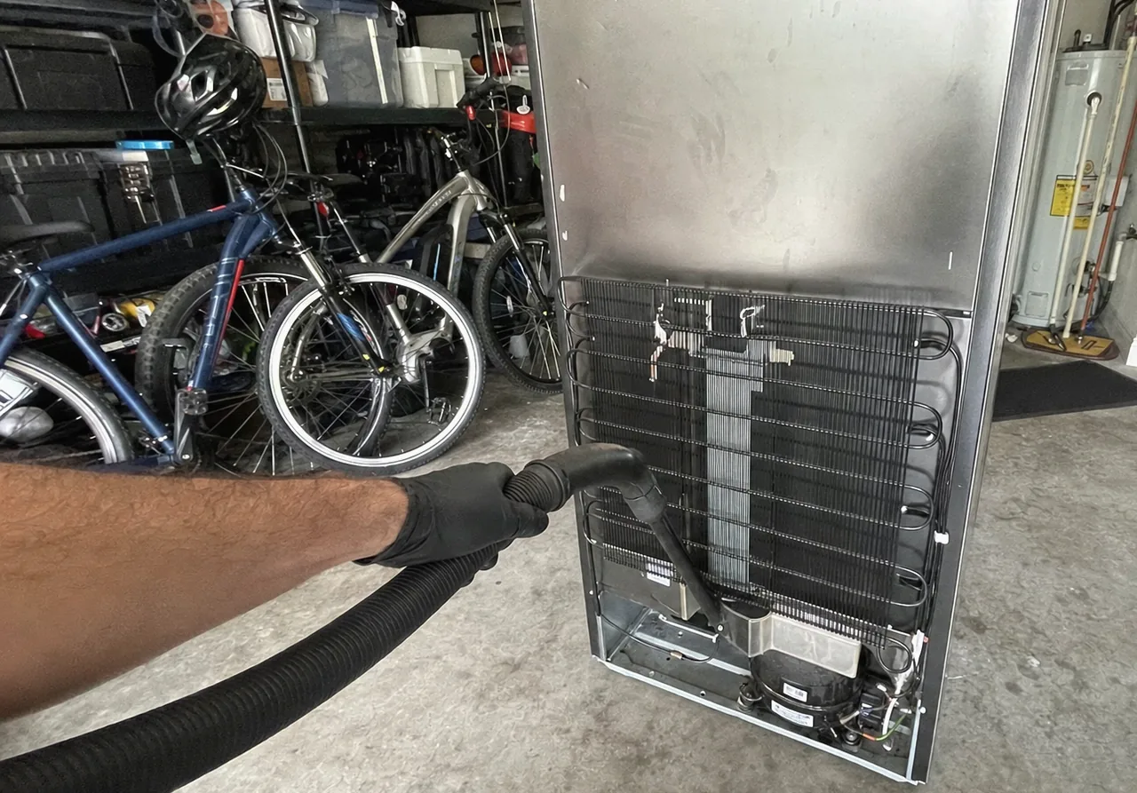 A refrigerator standing free of the wall with its rear panel off, the coil and compressor bay open to the garage floor