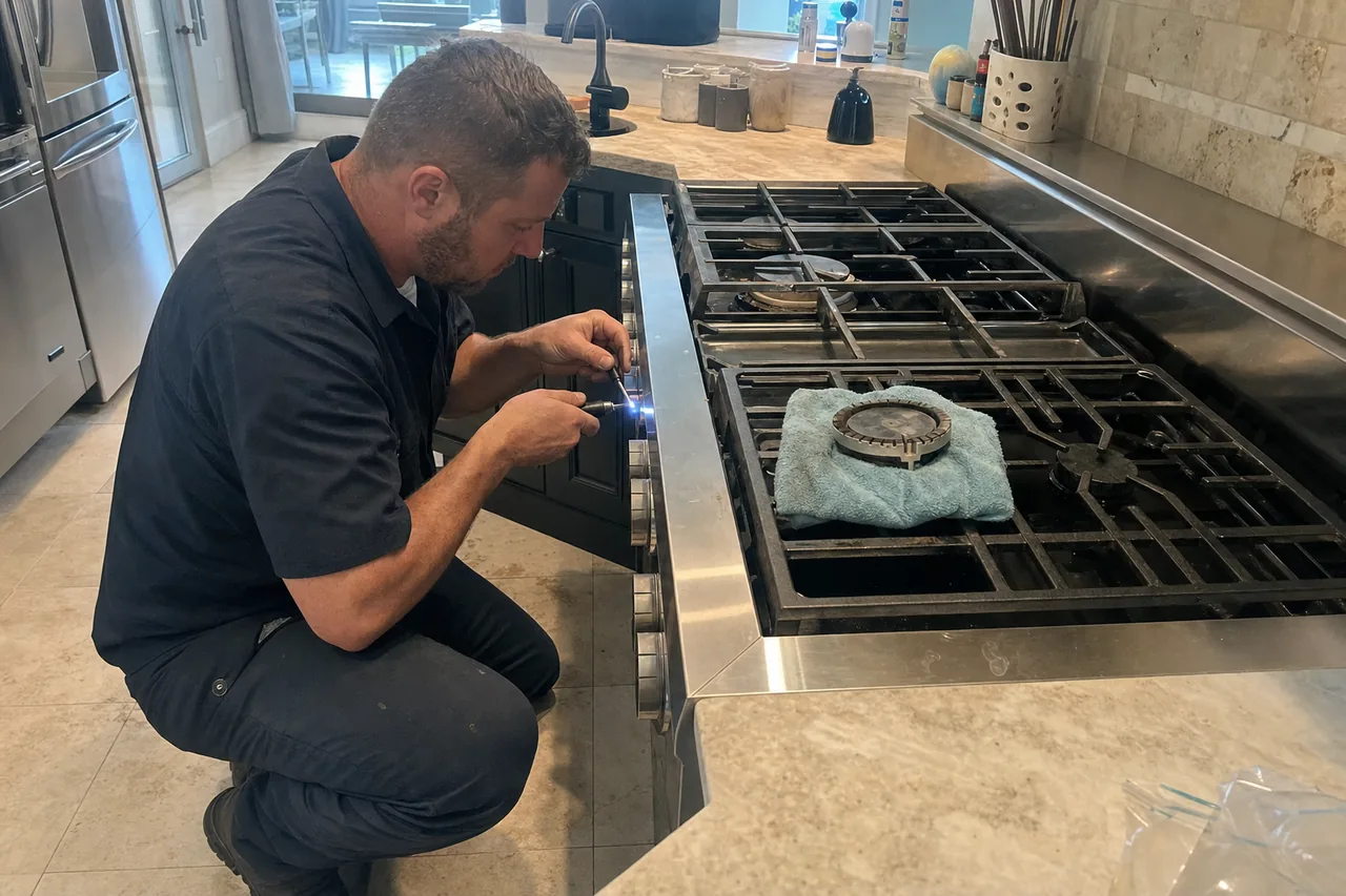 A technician crouched at the front of a stainless built-in range, working at burner height while one burner cap rests on a cloth on the grate