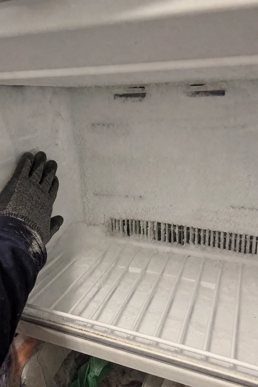 A freezer interior sheeted in frost from the ceiling down to the shelf, the vent slots at the back almost closed over with ice